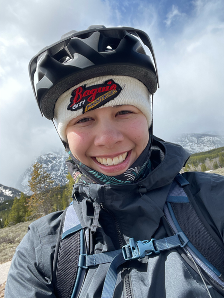 Bundled up smiling girl wearing a bike helmet with clouds and alpine scenery in the background.